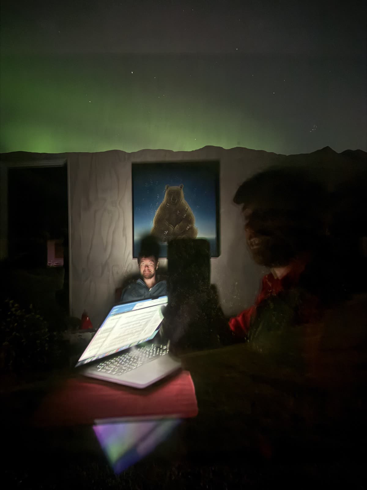 A dark room in Valdez at night. Two people at a
        table, one lit blue by a laptop screen, the other a large out-of-focus shape in the
        foreground. A painted bear hangs on the wall behind them, and the aurora is green
        over a mountain ridge through the window above.